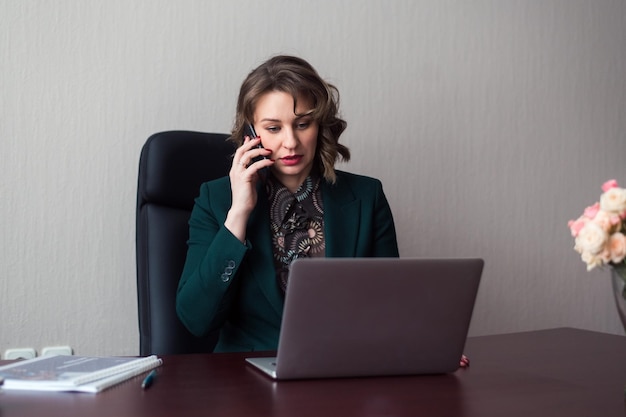 Young business woman or manager sitting at workplace with laptop and using smartphone