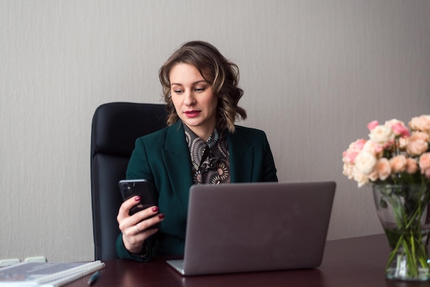 Young business woman or manager sitting at workplace with laptop and using smartphone