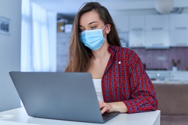 Young business woman in a medical protective mask works from home at the computer during self-isolation and quarantine.