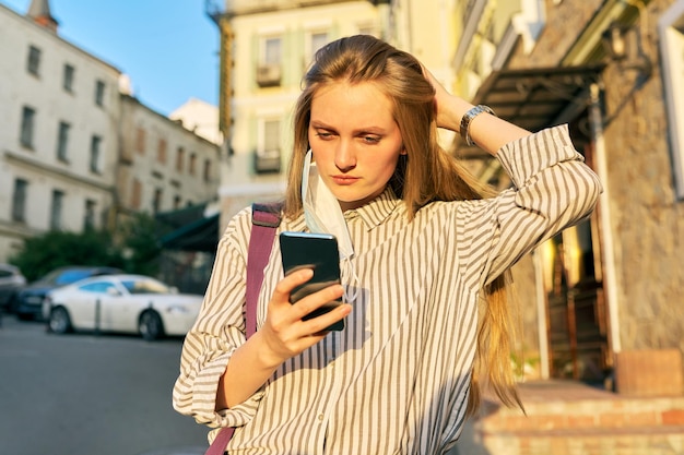 Young business woman office worker in medical protective mask with smartphone in hands, mask on ear open face. Female on street of city. Lifestyle, business in an epidemic, pandemic, bad ecology