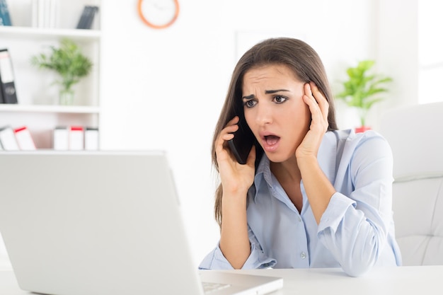 Giovane donna d'affari, telefonando in ufficio, con un'espressione preoccupata sul viso guardando il laptop che è di fronte a lei.