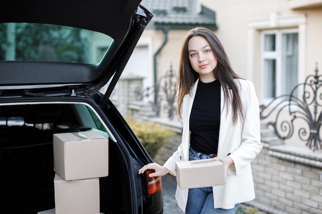 Молодая деловая женщина собирает посылки из багажника машины, возвращаясь домой на машине Концепция покупки товаров в Интернете и доставки их домой