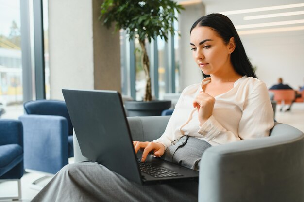 Молодая деловая женщина сидит на диване и работает с ноутбуком в зале ожидания бизнес-центра