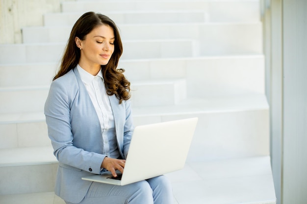 Молодая деловая женщина сидит на лестнице в коридоре офиса и работает на ноутбуке