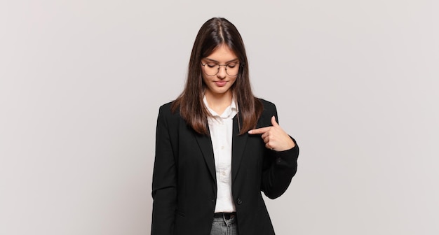 Young business woman smiling cheerfully and casually looking downwards and pointing to chest