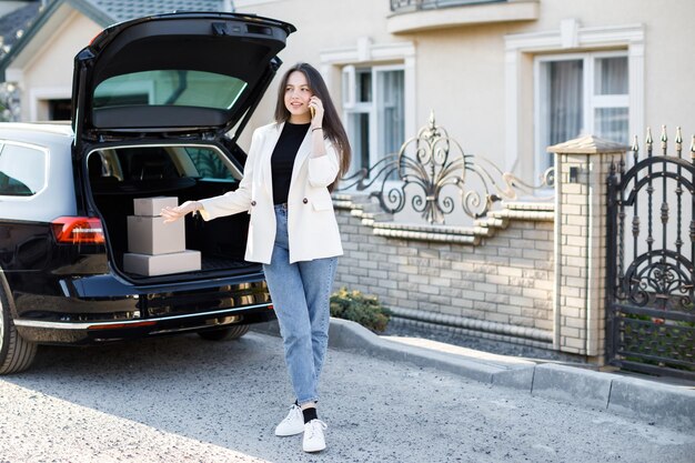 車で荷物が帰ってくる車の前に立っている若いビジネスウーマン女の子が車の近くに立って携帯電話を持っているオンラインで商品を購入して家に届けるというコンセプト