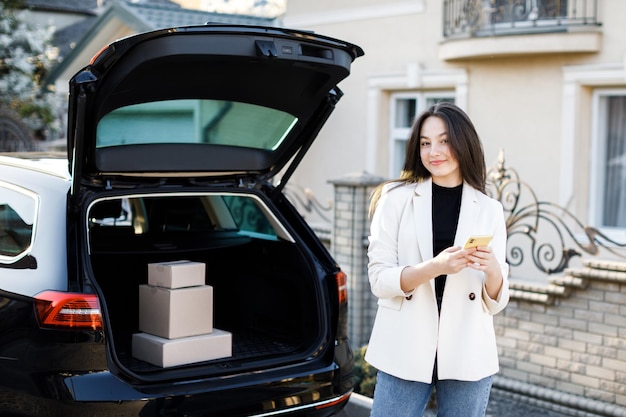 Young business woman standing at the car with parcels coming home by car The girl is standing near the car and holding a mobile phone Concept of buying goods online and delivering them home