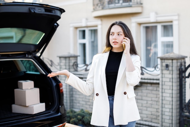 Young business woman standing at the car with parcels coming home by car The girl is standing and talking on mobile phone near the car Concept of buying goods online and delivering them home