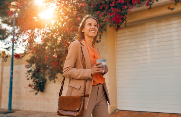 A young business woman or student in a suit smiles and drinks\
coffee while walking on the streets of a town full of flowers.