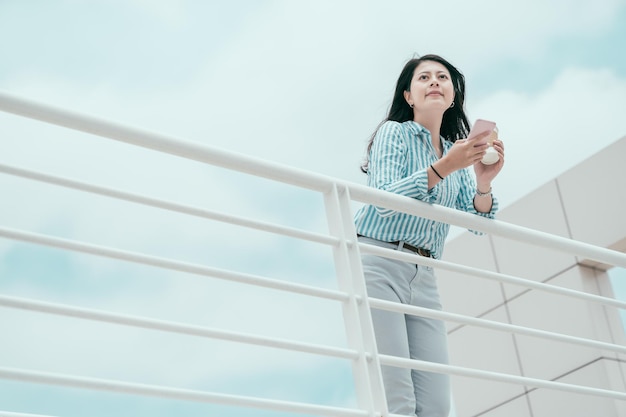 Young business woman texting message on her smartphone while leaning on railing during sunny day with blue sky. smiling and relaxed office lady enjoy sunshine using cellphone outside work building.