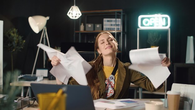 Фото Молодая деловая женщина разбрасывает бумаги в помещении в замедленной съемке удовлетворенная хипстерская девушка заканчивает проект в ночном офисе веселая сотрудница наслаждается результатами работы на рабочем месте