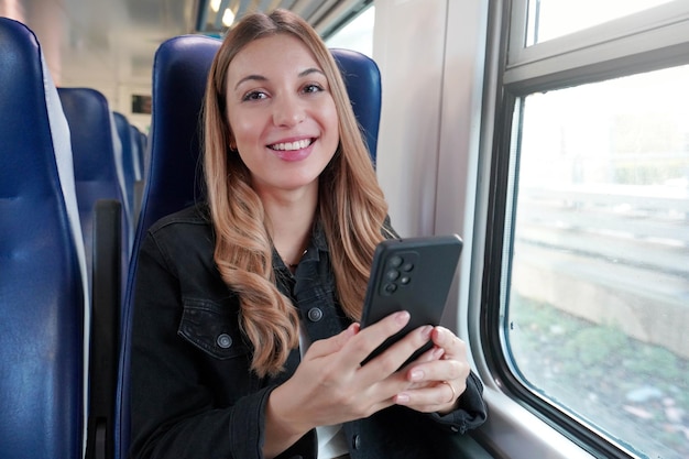 Молодая деловая женщина, использующая общественный транспорт, сидит с телефоном в поезде и смотрит в камеру