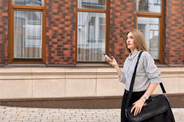 Young business woman walking in city talking on mobile phone profile view, office building background, copy space