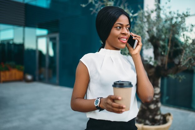 段ボールのコーヒーカップとオフィスビルに対して携帯電話を持つ若いビジネス女性