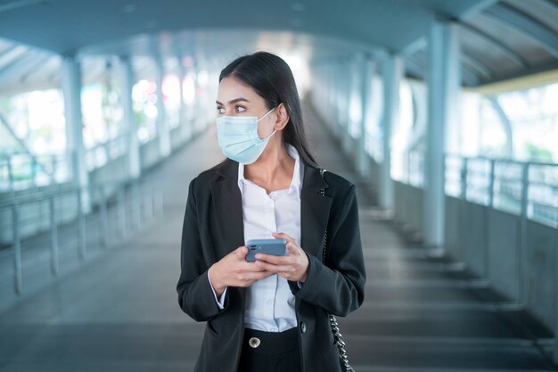 Молодая деловая женщина с маской для лица стоит на платформе метро, используя умный