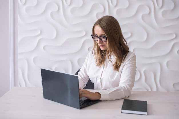 A young business woman with glasses is sitting at a white desk and looking at a laptop