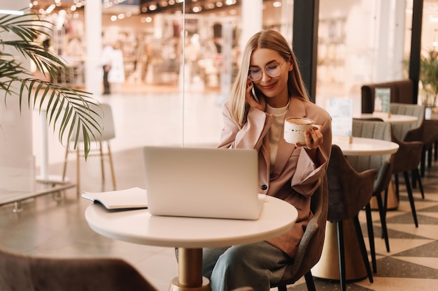 Una giovane donna d'affari con gli occhiali parla al telefono e lavora online utilizzando un laptop in un bar