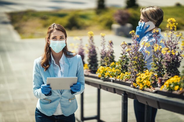 Молодая деловая женщина с защитной маской на лице что-то ищет на своем цифровом планшете на открытом воздухе во время пандемии COVID 19.