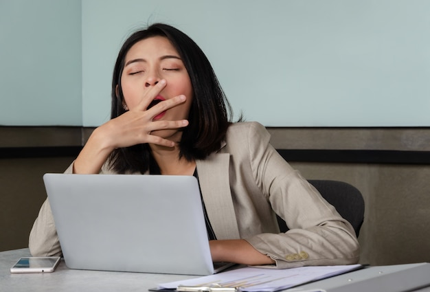 Young business woman yawning at meeting office table in front of laptop, covering her mouth out of courtesy. Overwork and sleep deprivation concept