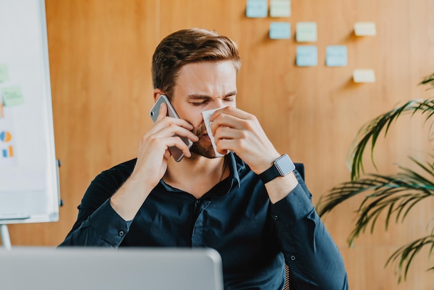 Young businessman blowing his nose with tissue at work while\
having conversation by mobile phone