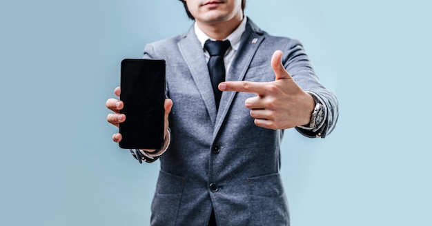 A young businessman brunette in a jacket, shirt and tie holds the phone and points his finger at the phone. Blank screen close up on the blue background