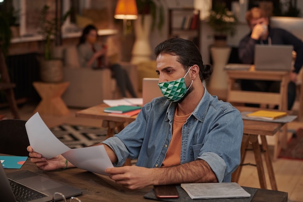 オフィスで書類と彼のテーブルで保護マスクで働くカジュアルな服装の青年実業家