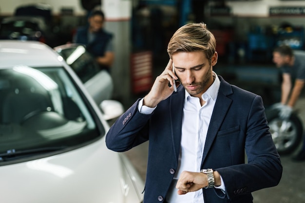 Young businessman checking the time on wristwatch while talking on mobile phone at auto repair shop