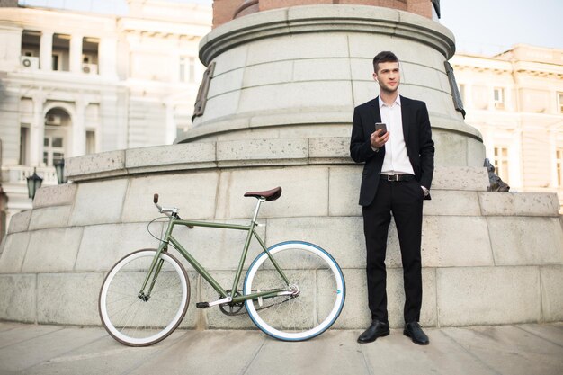 Молодой бизнесмен в классическом черном костюме и белой рубашке с беспроводными наушниками держит мобильный телефон в руке, задумчиво глядя в сторону с ретро-велосипедом рядом на улице