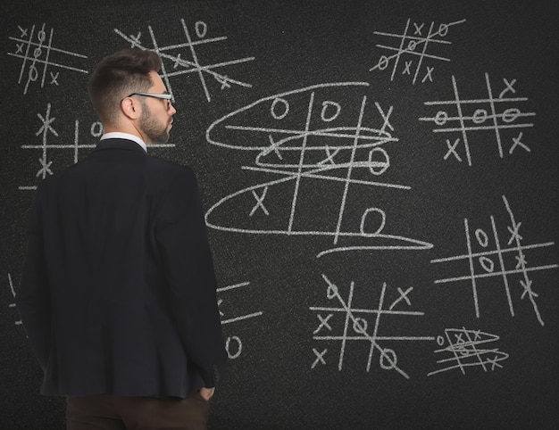 Young businessman in elegant suit near blackboard with drawn tic tac toe game