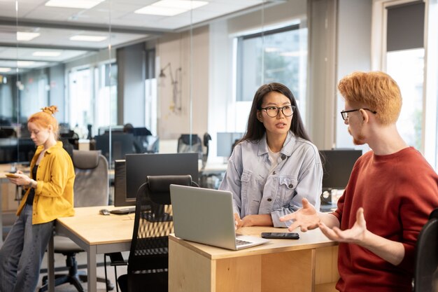 Young businessman in eyeglasses and casualwear discussing new ideas for project with Asian female colleague in front of laptop at working meeting