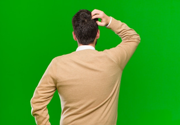 Foto giovane uomo d'affari sentirsi all'oscuro e confuso, pensando a una soluzione, con la mano sull'anca e altri sulla testa, vista posteriore sul muro verde