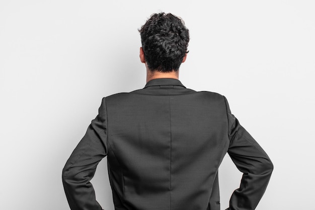 Young businessman feeling confused or full or doubts and questions, wondering, with hands on hips, rear view