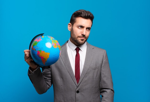 Photo young businessman feeling sad, upset or angry and looking to the side with a negative attitude, frowning in disagreement holding a world globe map