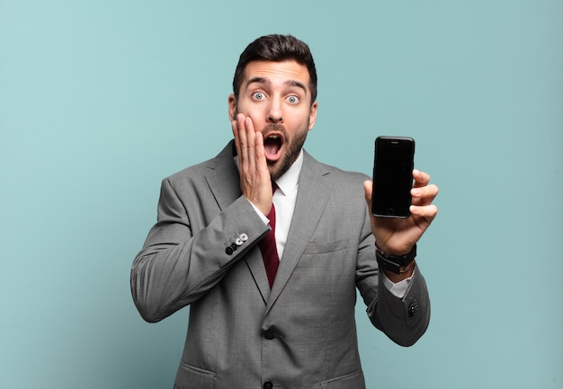 Young businessman feeling shocked and scared, looking terrified with open mouth and hands on cheeks and showing his phone screen