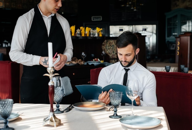 Un giovane uomo d'affari in un raffinato ristorante esamina il menu e fa un ordine a un giovane cameriere con un elegante grembiule servizio clienti servizio al tavolo nel ristorante