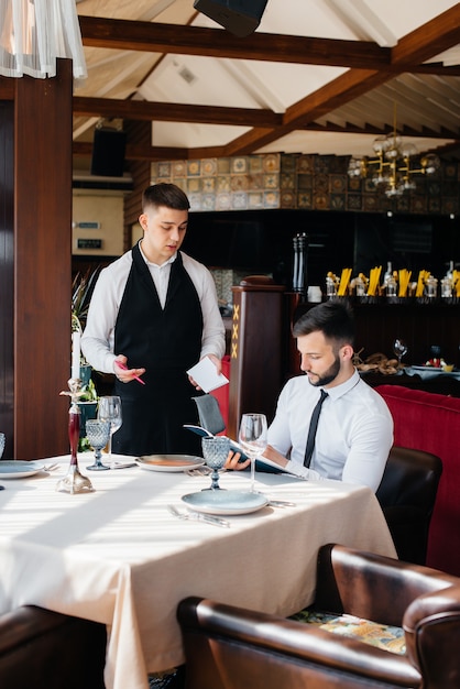 A young businessman in a fine restaurant examines the menu and makes an order to a young waiter in a stylish apron. Customer service.