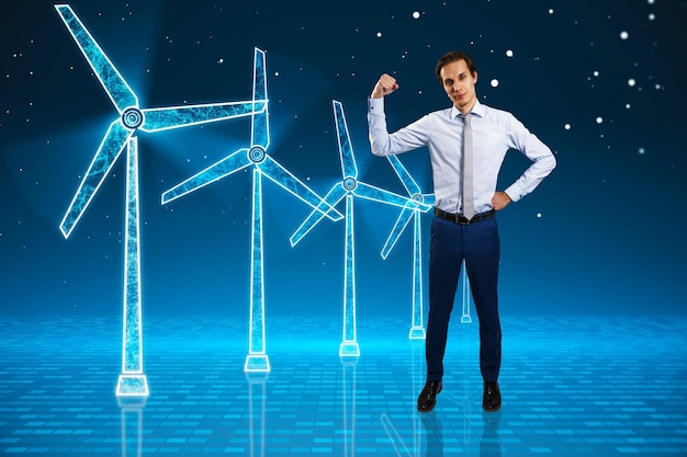 Young businessman flexing muscle and looking at glowing digital wind mill turbine hologram on blue background Wind generator concept