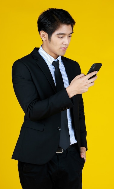 Young businessman in formal black suit. Standing look at mobile phone seems texting message. Yellow background gives modern feel. The space can fill with text.