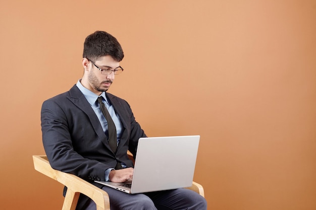 Young businessman in glasses concentrated on work sitting in armchair and typing on laptop