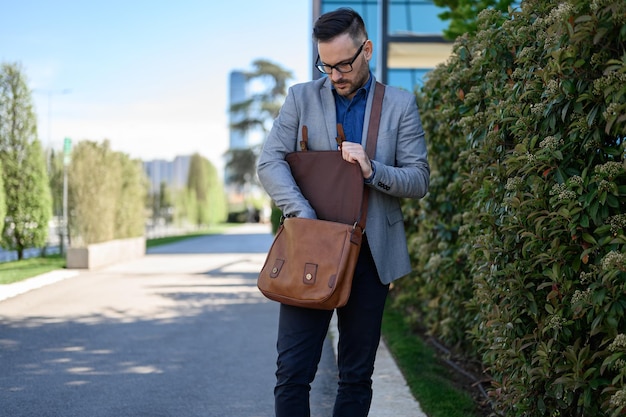 young businessman in glasses searching brown laptop bag while standing by plants on city street