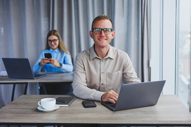 A young businessman is looking at something on a laptop while\
working the concept of a modern successful person young confused\
guy in glasses sitting at desk in open space office with\
laptops