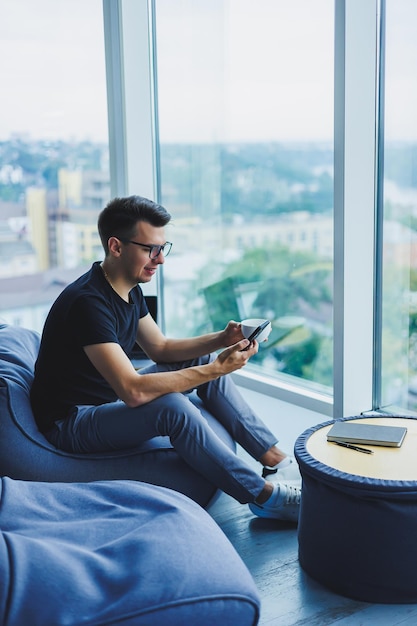 A young businessman is looking for something and typing on a smartphone and drinking fresh black coffee The concept of a modern successful person Young focused guy with glasses in an open office