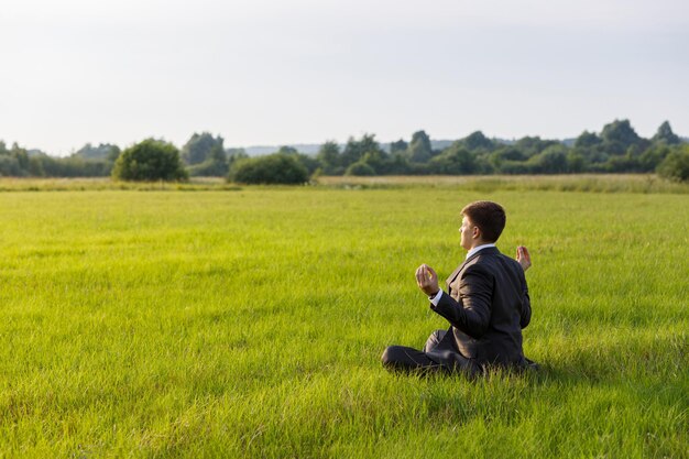 Young businessman is meditating sitting on the grass in a field