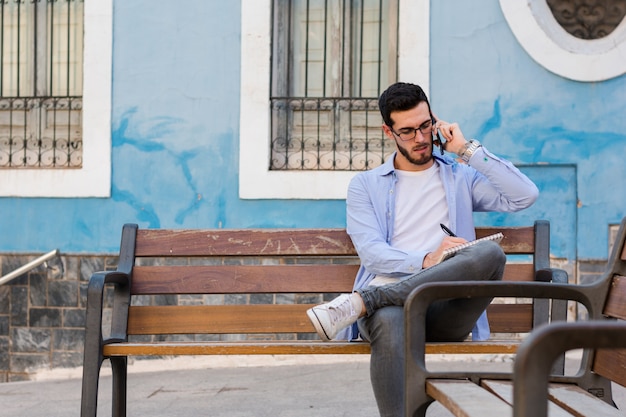 Молодой бизнесмен сидит на скамейке, в то время как он разговаривает по мобильному телефону и пишет в своей записной книжке