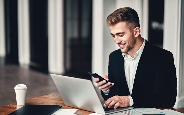 A young businessman is sitting in a business center with a laptop and a phone in his hand, on the table are documents and coffee