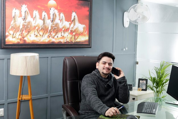 A young businessman is sitting in the office talking on mobile and smiling.