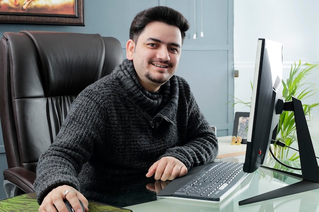 A young businessman is sitting in the office working on computer and smiling