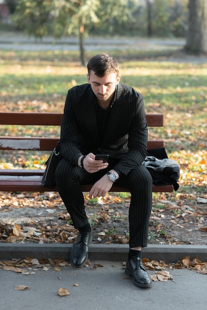 A young businessman is sitting on a park bench using a smartphone