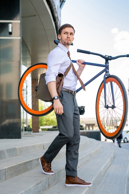 仕事の後にオフィスビルを離れ、都市環境で階下に行く間彼の自転車を運ぶ青年実業家