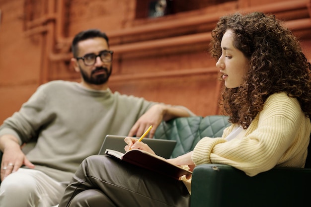 Young businessman listening to his female colleague making notes in notebook